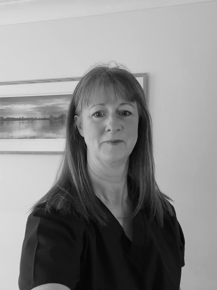 A black and white headshot portrait of a red-haired woman wearing a blue v-neck short-sleeved shirt, stood in front of a cream coloured wall with a framed photo hung on it.