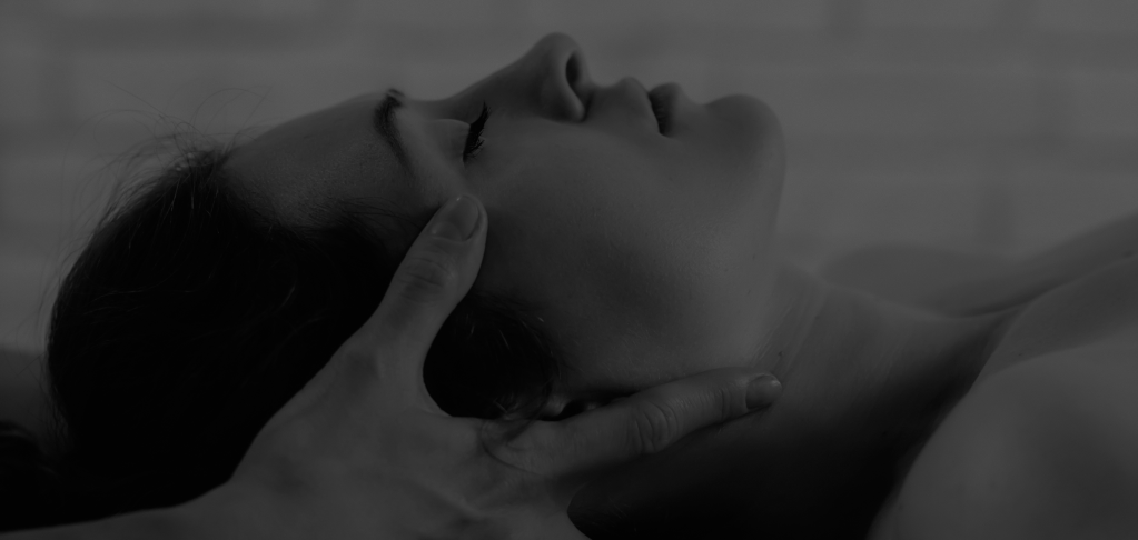 A low brightness monochrome image of a woman shown from a side angle as she lies down, while someone elses hands are placed on the sides of her head.